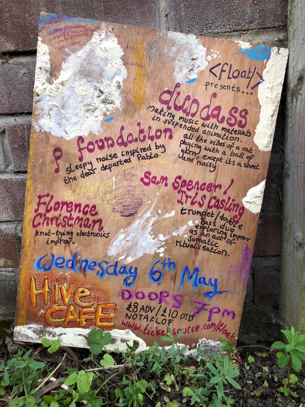 Gig poster containing the event details listed below, text appears to be doodled on a piece of plywood with paint splashed on it, leant against a brick wall on a concrete path with weeds coming through the cracks.