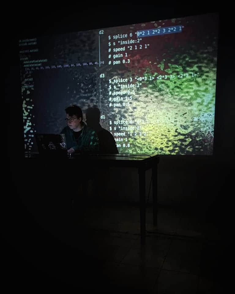 Charlotte sat at a desk performing with a laptop. Behind her is a projection of code overlayed on a purple, green and white fluid like texture.